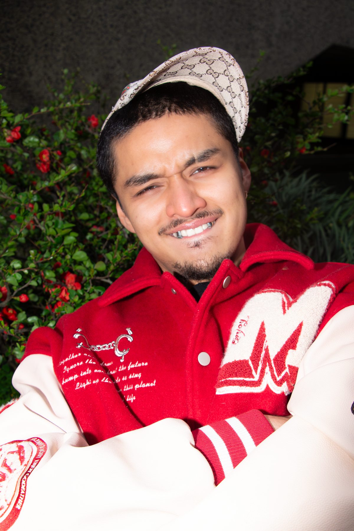 Ruben in a red varsity jacket and monogram cap grins to show silver caps, red flowers behind