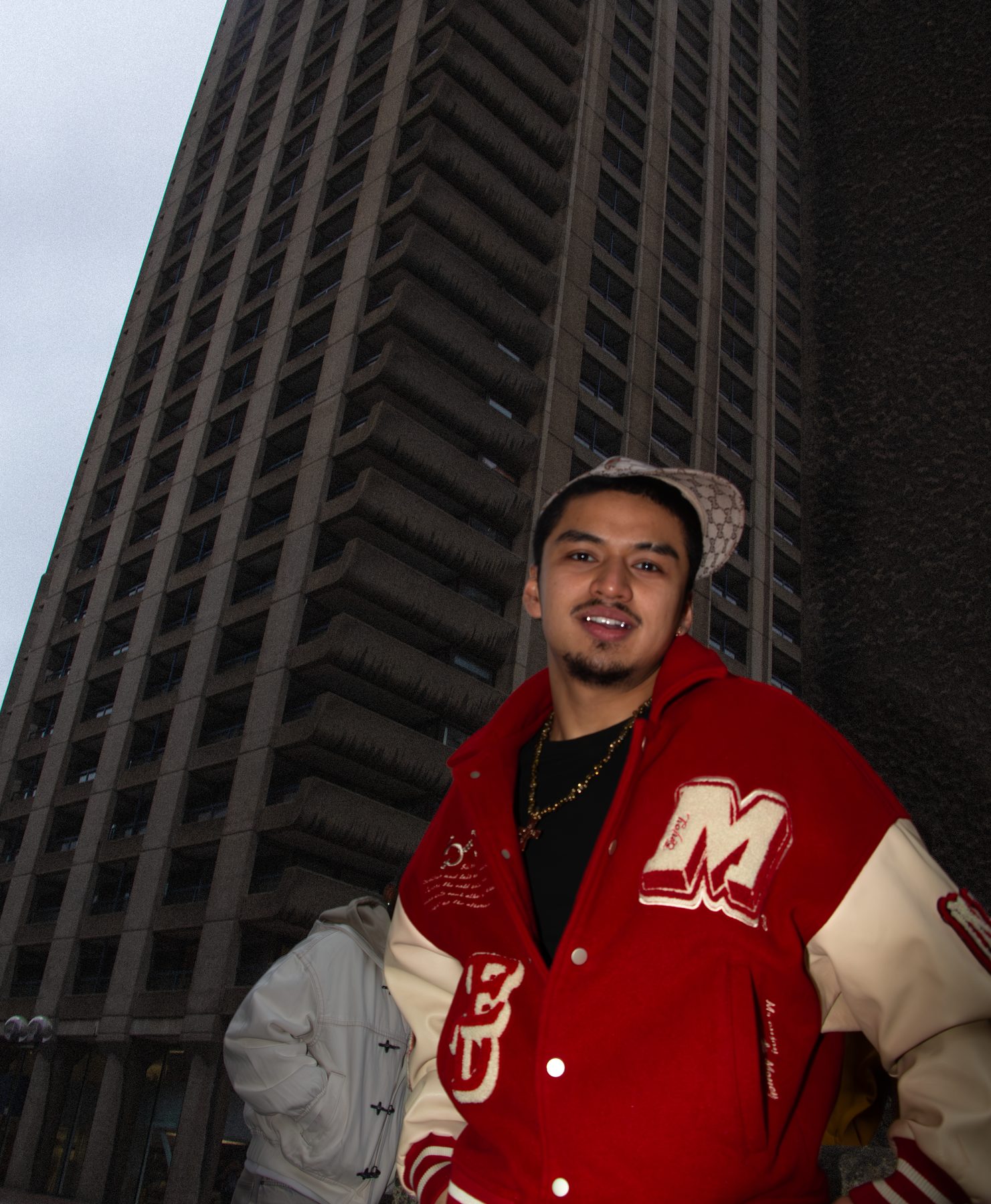 Ruben in a red varsity jacket stands below a Barbican tower block, smiling