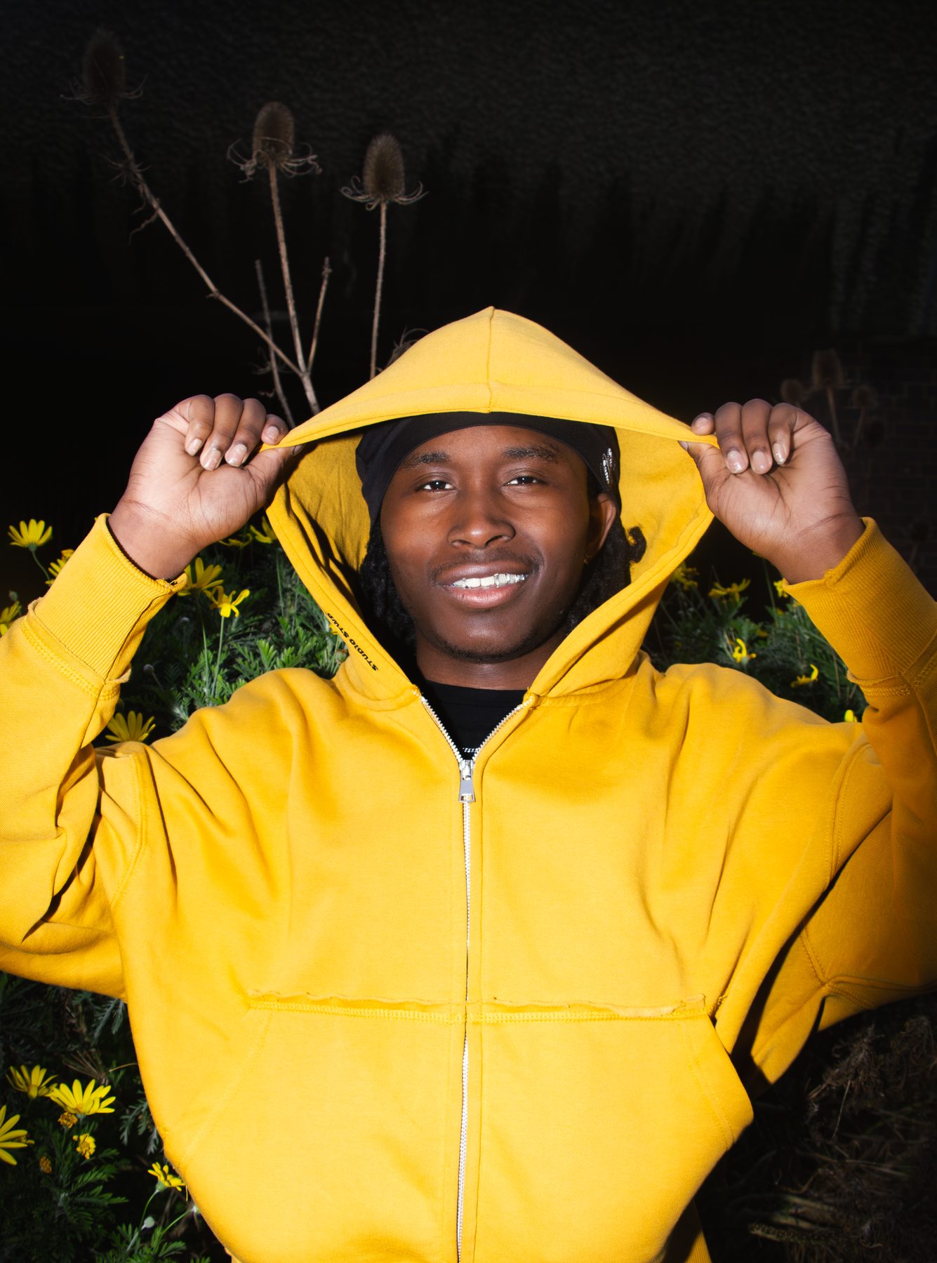 Shaq holds up a yellow hood at night, smiling to show silver grillz among yellow flowers
