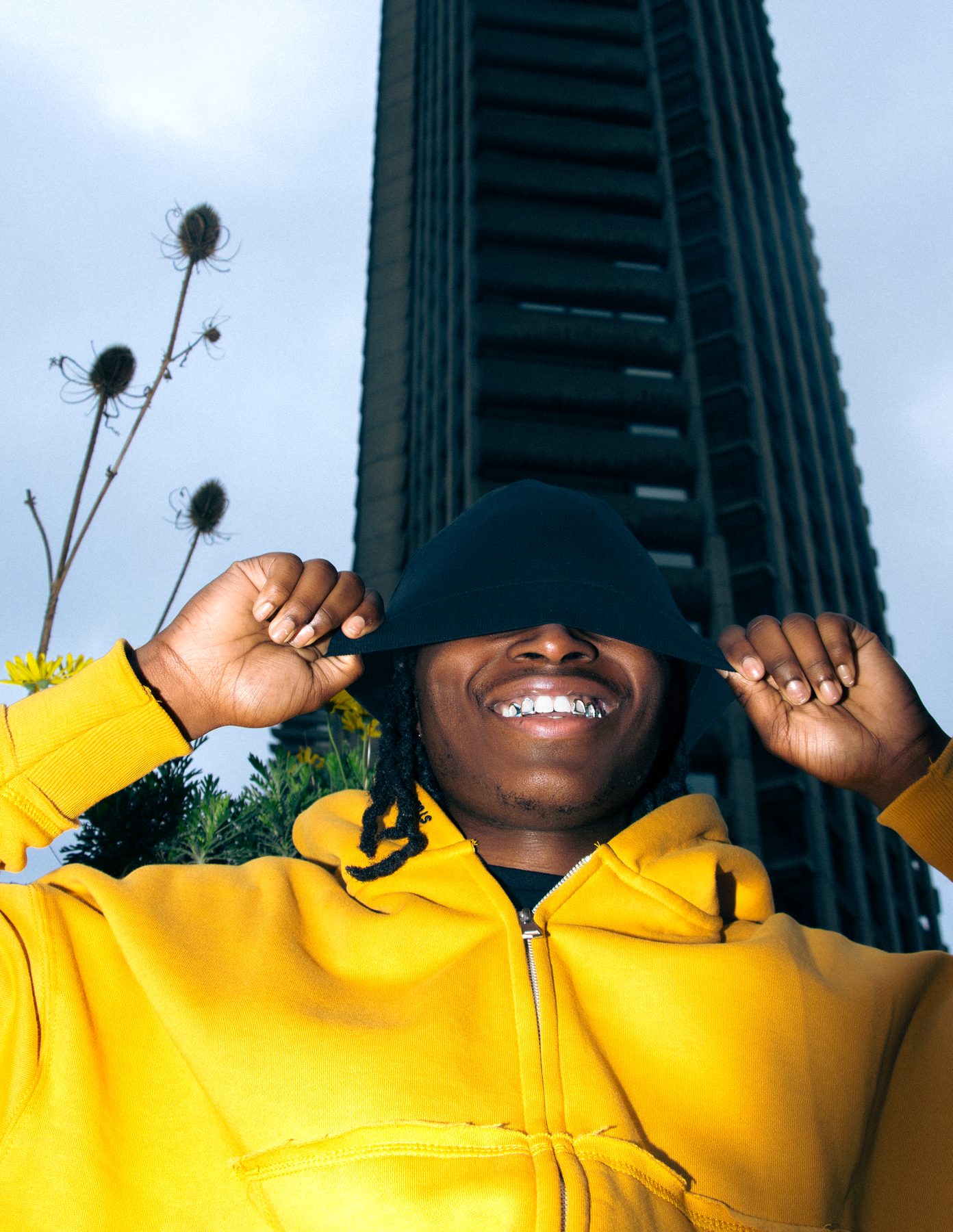 Shaq in a yellow hoodie pulls a black hood over his eyes, smiling to show silver grillz, beneath a Barbican tower